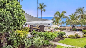 A covered outdoor seating area with a view of the ocean, surrounded by palm trees and lush greenery. - 🏖️Garden View Kona Condo | A/C, Pools, Walk to Ocean (Kailua Kona)
