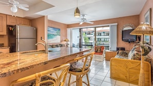 A modern kitchen with a large island, bar stools, and a dining area with wicker furniture. - 🏖️Garden View Kona Condo | A/C, Pools, Walk to Ocean (Kailua Kona)