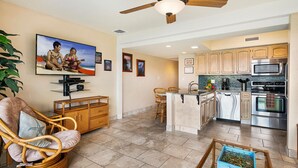 A living room with a television, a wicker chair, and a kitchen with modern appliances. - 🏖️Kona Oceanview Condo w/ Lanai + A/C | Casa 328 (Kailua Kona)