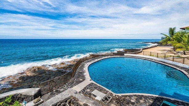 A pool with a curved edge, surrounded by a stone wall and a railing, overlooking the ocean. - 🏖️Kona Oceanview Condo w/ Lanai + A/C | Casa 328 (Kailua Kona)