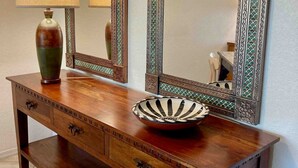 Um console de madeira com duas gavetas, um vaso e uma tigela. Dois espelhos com molduras decorativas estão fixados na parede acima do móvel.