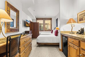 A hotel room with a bed, a dresser, a desk, a chair, a desk lamp, a wall-mounted TV, a window with blinds, and a door. - Main Street Condo|Steps to Lift |Amazing Rooftop I Managed by Park City Lodging (PARK CITY)