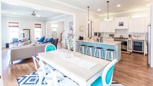 A modern kitchen with white cabinets, a central island with stools, and a dining area with a table and chairs. - Carlton Landing! Beautiful home with screened porch and outdoor fireplace! (Carlton Landing)