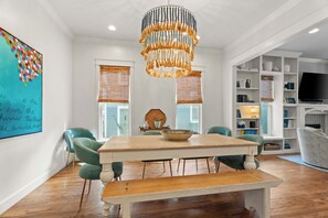 Een eetruimte met een houten tafel, groene stoelen, een bank, een decoratieve kroonluchter en een boekenkast met een televisie.