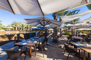 Area pranzo all'aperto con tavoli, sedie e ombrelloni, con una piscina sullo sfondo.