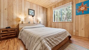 Une chambre à coucher aux murs lambrissés, avec un lit, des lampes de chevet, une table de chevet et une fenêtre donnant sur des arbres.