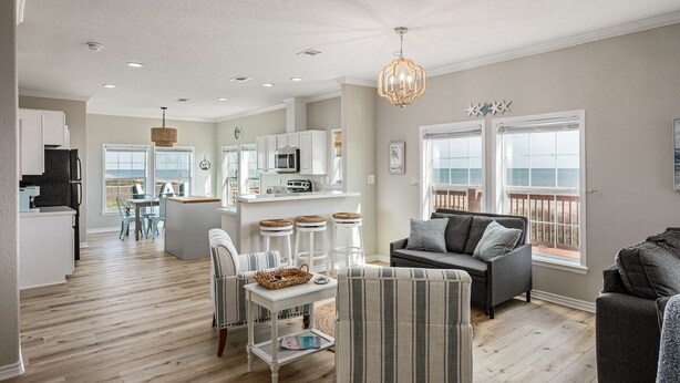 A spacious living area with a gray sofa, a striped armchair, and a dining table with stools. There is a kitchen area with white cabinets and a microwave. The room has large windows with a view of the ocean.