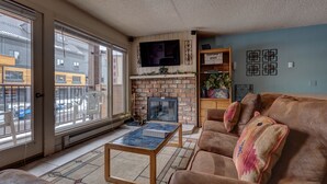 Un salon doté d’une cheminée, d’un téléviseur fixé au-dessus, d’un canapé agrémenté de coussins décoratifs et offrant une vue sur l’extérieur grâce à de grandes fenêtres.