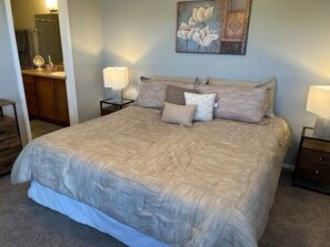 Un lit bien fait, avec une couette beige et plusieurs oreillers, une table de chevet avec une lampe et un tableau accroché au mur.
