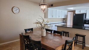 Un coin repas avec une table en bois, des chaises noires, un vase avec des fleurs, une horloge murale, et une cuisine en arrière-plan.