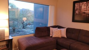 A living room with a brown sectional sofa, a small table with a lamp, and a large window with a view of trees and mountains. - Amazing Mountain Views from this 2nd Floor Large 2 Bedroom in Ventana Canyon! (Tucson)