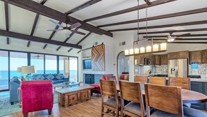 A spacious living area with a large window overlooking the ocean, wooden dining table, and a red sofa.