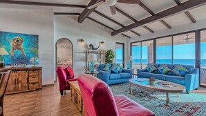 A spacious living room with a blue sofa, red armchair, and a coffee table. There is a wooden cabinet and a painting on the wall.