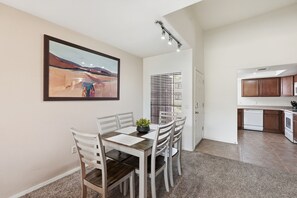 Un coin repas avec une table et des chaises, un tableau accroché au mur, une cuisine équipée de placards et d’appareils électroménagers, et une fenêtre avec des stores.