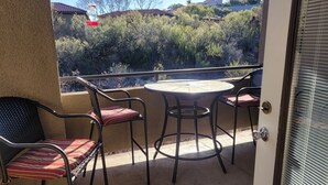 A balcony with a round table and four chairs, a door with a window, and a view of a house and trees. - Tucson flair in this 2nd floor-serene patio -close to main pool- exercise rm (Tucson)