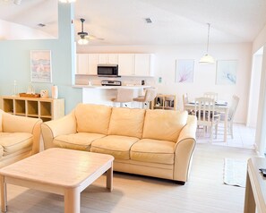 A living room with a beige sofa, a wooden coffee table, and a kitchen area with white cabinets and a microwave.