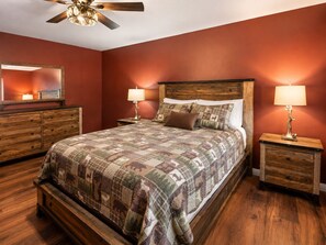 Une chambre à coucher avec un grand lit, une tête de lit en bois, deux tables de chevet avec des lampes, une commode en bois et un ventilateur de plafond.