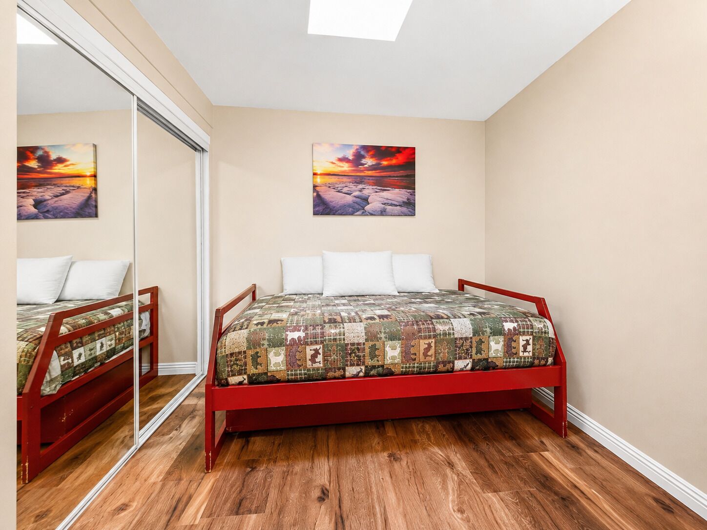 A bedroom with a wooden floor, a red bed frame, a quilted bedspread, and two wall-mounted paintings.