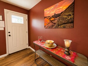 A room with a wooden floor, a white door, a wall-mounted painting of a sunset, and a table with a candle and a bowl.