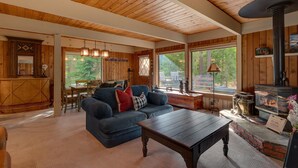 A cozy cabin interior with a wooden ceiling, a fireplace, a dining area, and large windows offering a view of the outdoors. - Cozy Wood Cabin By Tahoe Getaways - 3 BR w Hot Tub, Fireplace + Backs to Forest (Carnelian Bay)