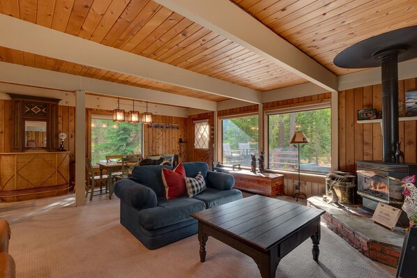 L’intérieur chaleureux d’un chalet avec un plafond en bois, une cheminée, un coin repas et de grandes fenêtres donnant sur le paysage extérieur.
