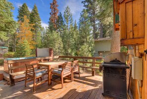 Une terrasse en bois avec un jacuzzi, un barbecue et un espace salon extérieur, le tout entouré d’arbres.