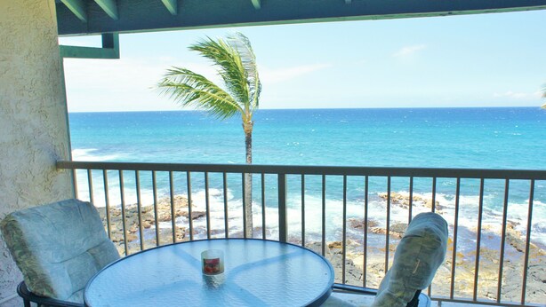 Un balcon avec une table ronde et deux chaises, donnant sur une plage avec un palmier et l’océan.