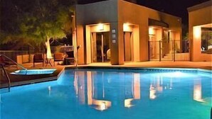 Una piscina con superficie de agua azul cristalina, rodeada por un edificio con varias puertas y ventanas, y un árbol a la izquierda.