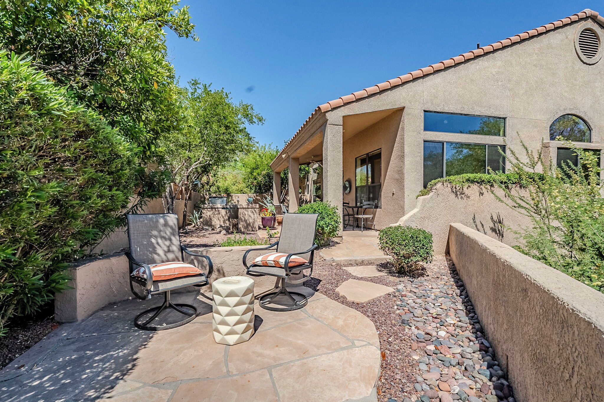 A patio area with outdoor furniture, including two chairs and a table, surrounded by a stone pathway and greenery.
