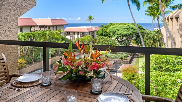Ein Holztisch mit tropischer Blumenarrangements, ein Glas Wasser und ein Teller auf einem Balkon mit Blick auf einen Resort mit Palmen und Meerblick.