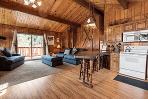 A cozy cabin interior with a wooden ceiling, a dining area with a bar, a kitchen with a stove, and a large window overlooking a deck.
