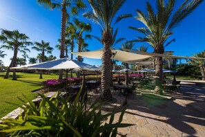An outdoor patio area with tables and chairs under large umbrellas, surrounded by palm trees and a well-maintained lawn. - Resort Living in Ocean View Penthouse at Esperanza (Cabo San Lucas)