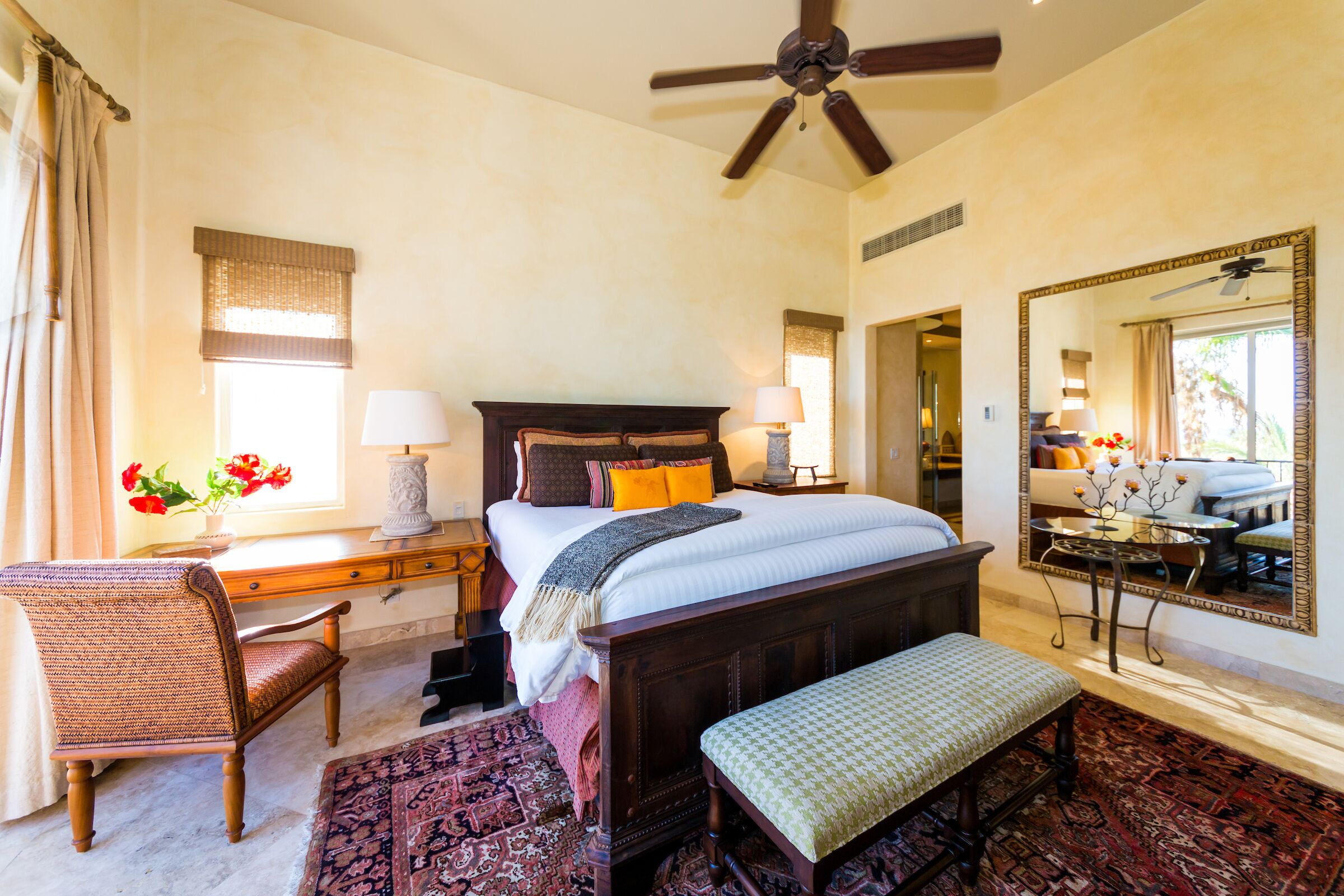 A bedroom with a large bed, a desk, a chair, a bench, a ceiling fan, and a mirror.
