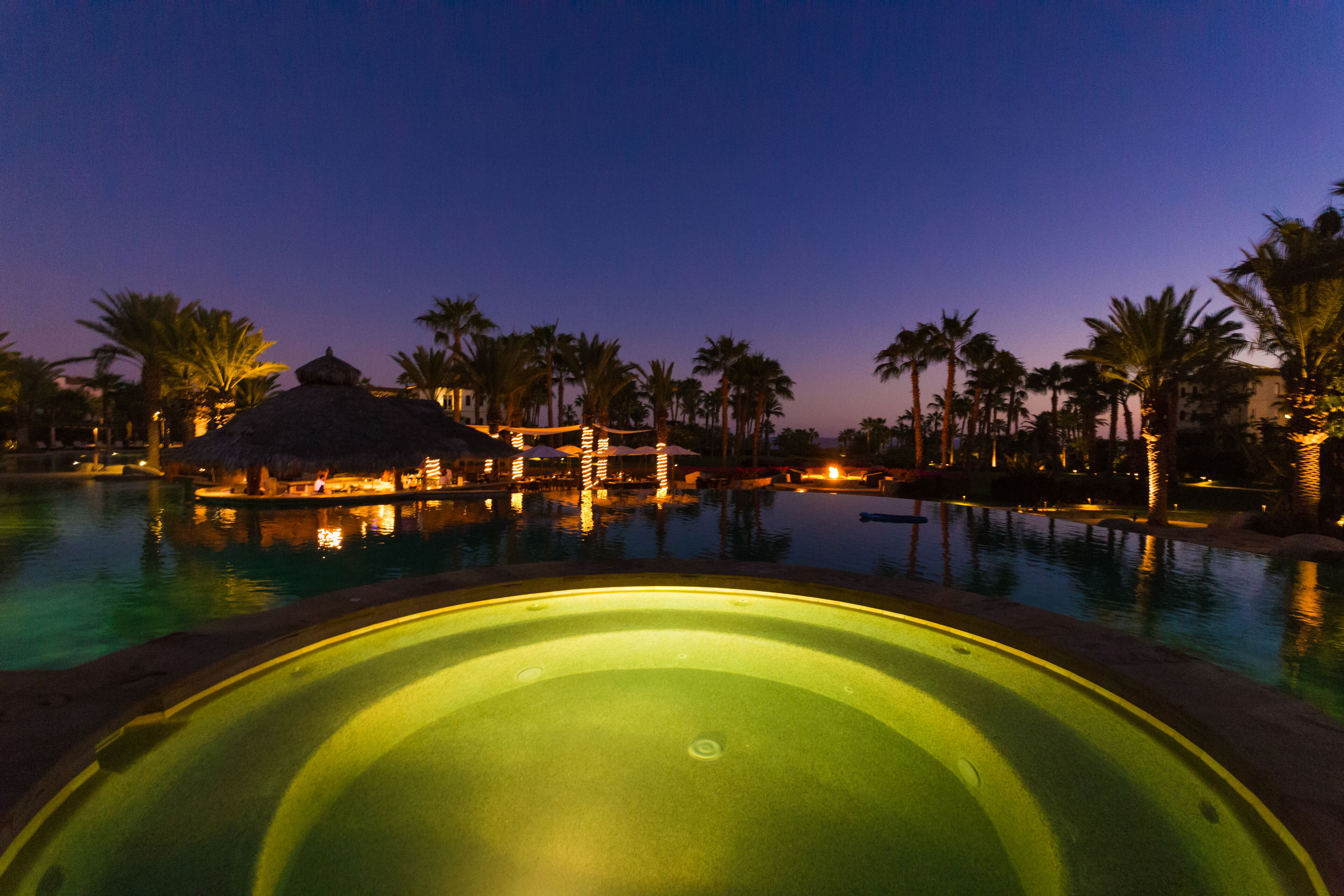 Ein Pool in einem Resort bei Nacht, mit Palmen und einer Struktur mit Reetdach.