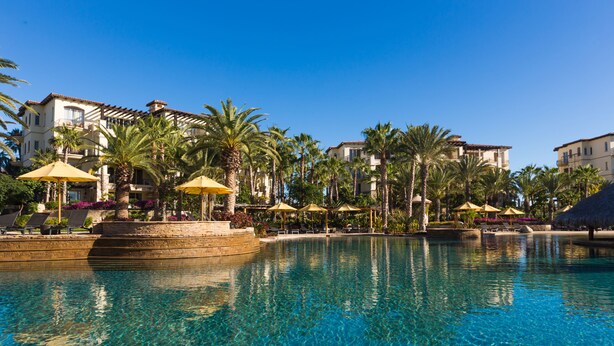 A clear blue pool with lounge chairs and umbrellas, surrounded by palm trees and buildings. - Resort Living in Ocean View Penthouse at Esperanza (Cabo San Lucas)
