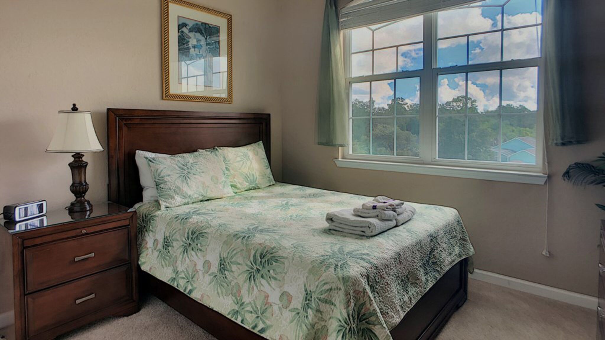 A bedroom with a bed, a nightstand, a lamp, a framed picture, and a window with a view of trees and a house.