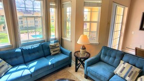 A living room with two blue sofas, a small round table with a lamp, and large windows offering a view of a pool and buildings. - Elegant Condo w/Pool View Balcony Resort Pool & Gym. Near Disney/Shops (Davenport)