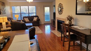 Un salon comprenant un canapé, une table basse et une table à manger. On y trouve également une cheminée, un ventilateur de plafond et une fenêtre avec des stores.