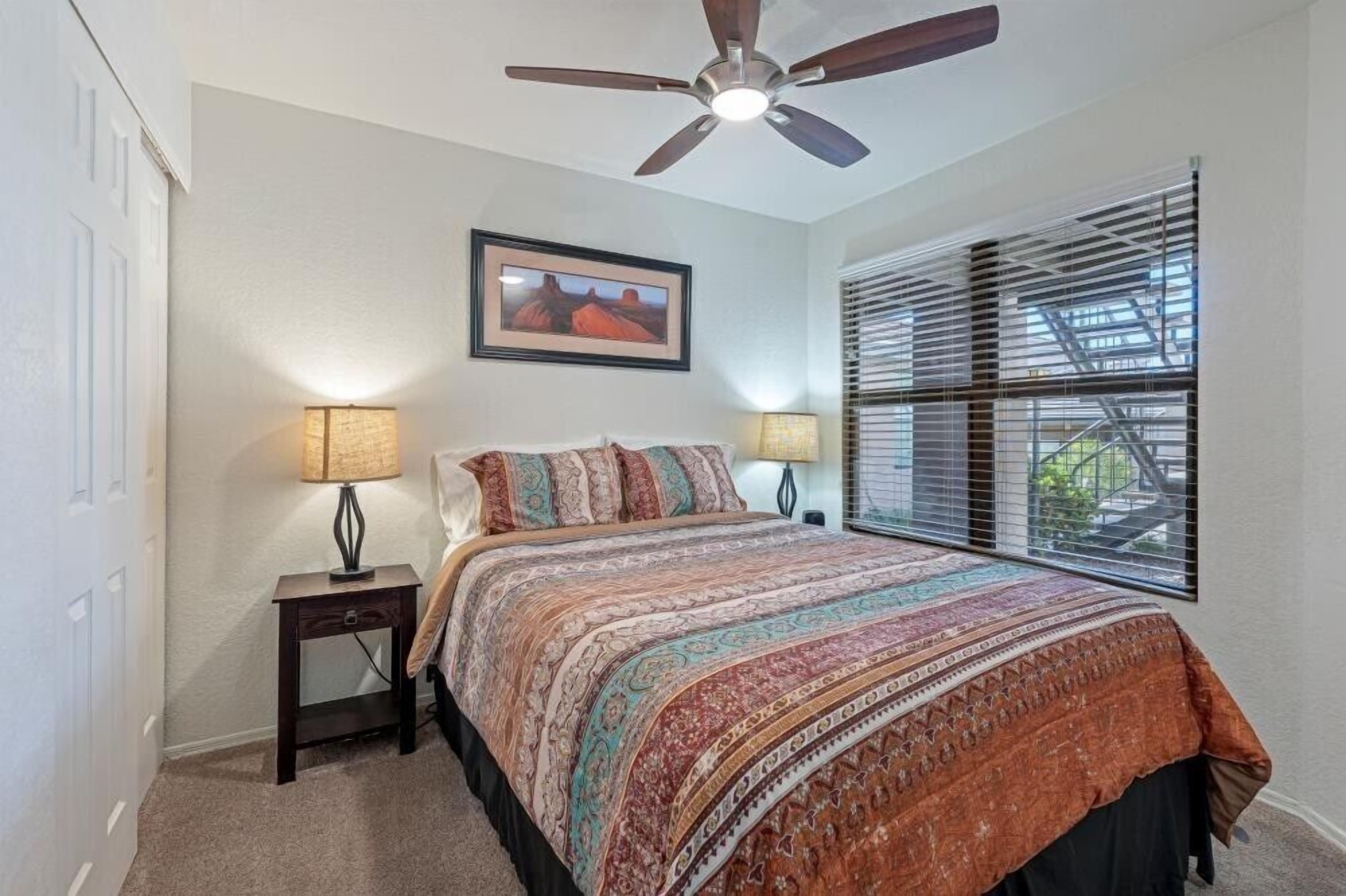 A bedroom with a bed, two bedside lamps, a ceiling fan, a framed picture on the wall, and a window with blinds.