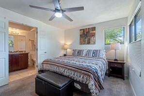 Une chambre à coucher avec un lit, un ventilateur de plafond, une table de chevet, une lampe, une fenêtre et une salle de bain visible à travers une porte ouverte.
