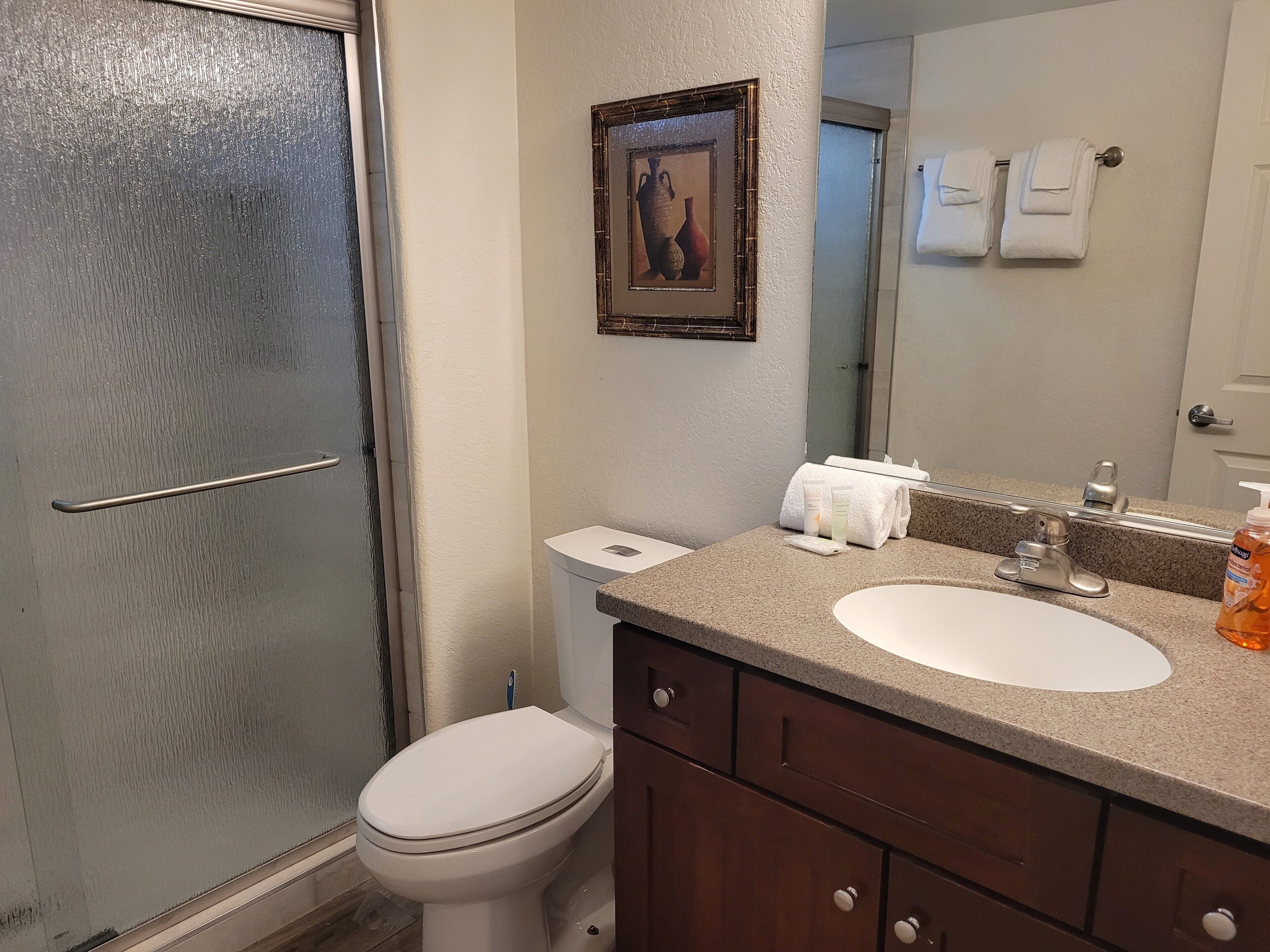 A bathroom with a toilet, sink, and shower. There is a framed picture on the wall, a towel rack with towels, and a bottle of hand soap on the sink counter.