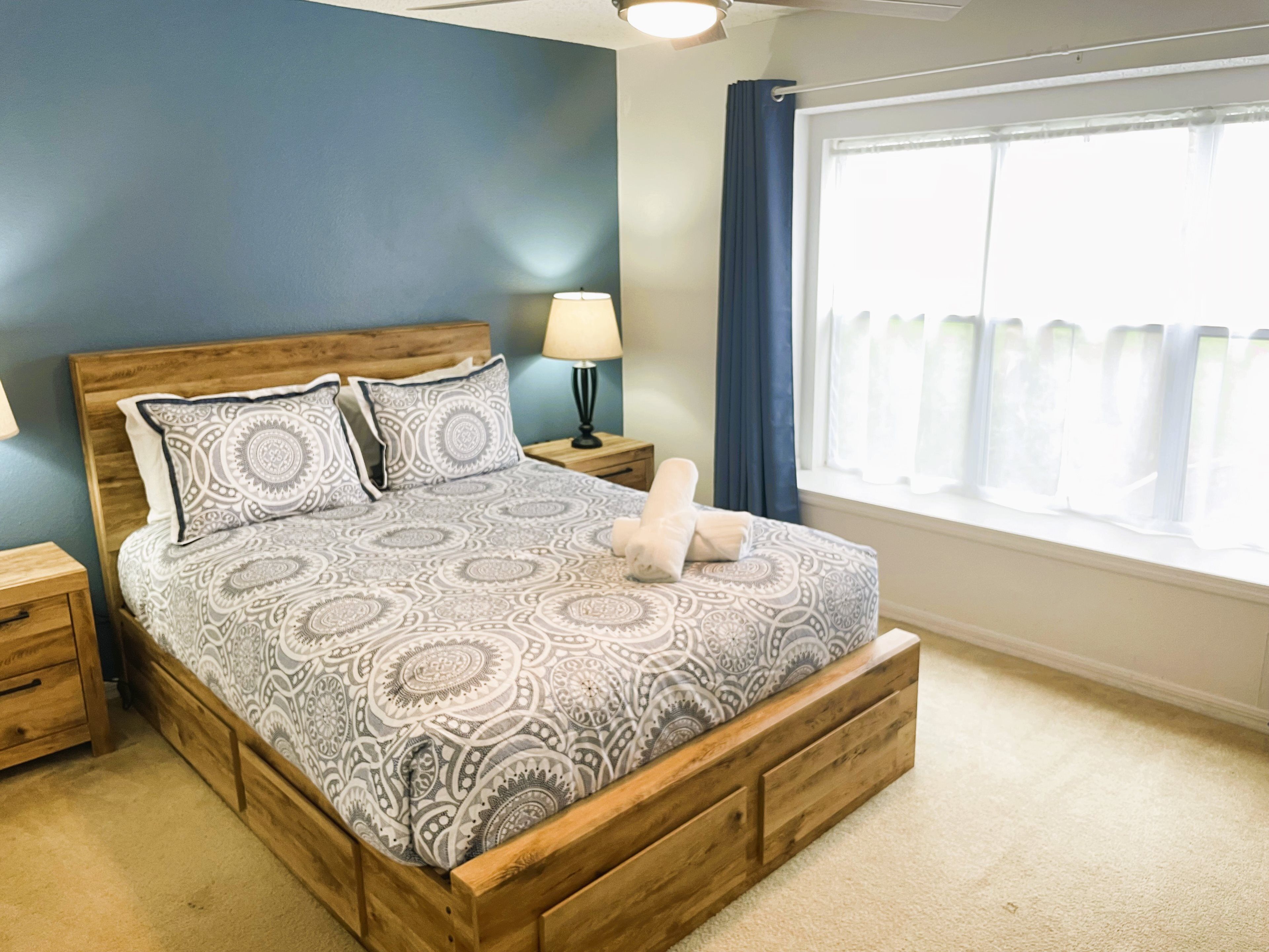 A bedroom with a wooden bed frame, patterned bedding, a nightstand with a lamp, a window with curtains, and a carpeted floor.
