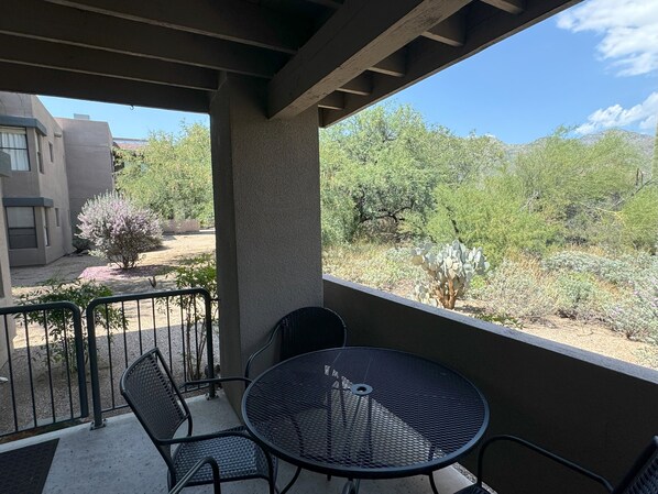 A balcony with a round metal table and chairs, overlooking a desert landscape with sparse vegetation and a building in the background.