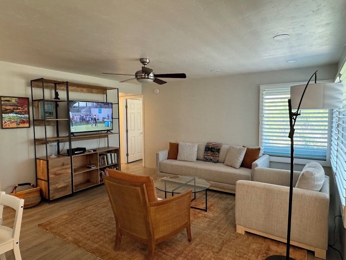 A living room with a sofa, armchairs, a coffee table, a TV on a wooden shelf, and a ceiling fan.