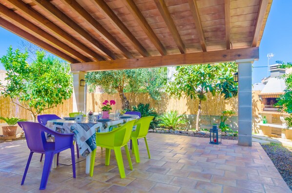 An outdoor patio with a wooden pergola, a table set for dining, and colorful chairs.