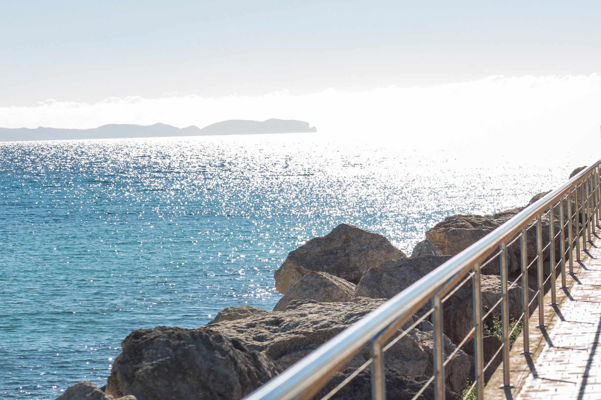 A coastal walkway with a metal railing, rocky terrain, and a view of the sea.