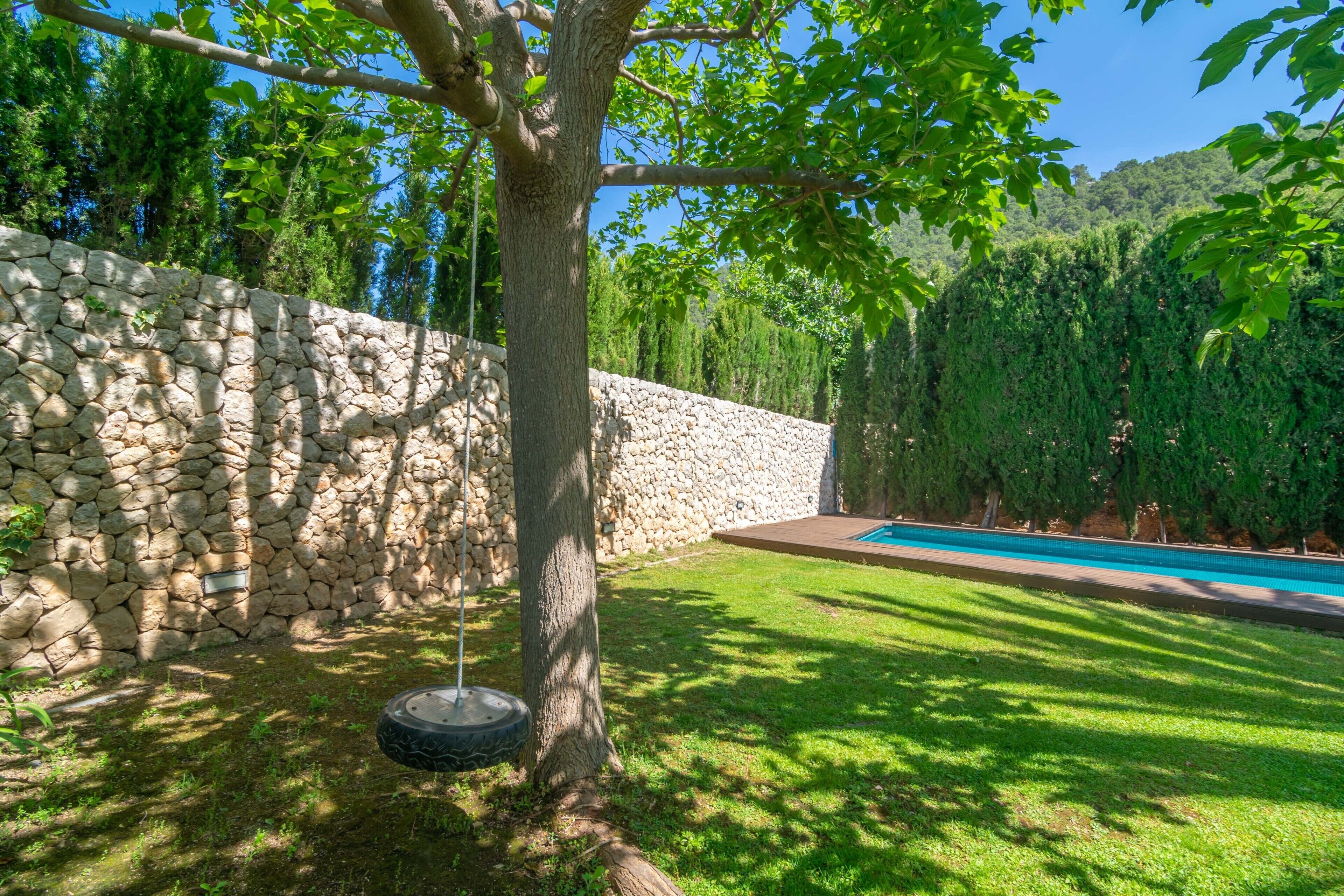 Un mur de pierre, un arbre, une balançoire, une zone herbeuse et une piscine.