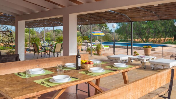 Ein Essbereich im Freien mit einem langen Holztisch, der für eine Mahlzeit gedeckt ist, umgeben von Stühlen und einem Pool im Hintergrund.