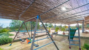 Ein Spielplatz im Freien mit Schaukeln, einer Rutsche und einer Kletterkonstruktion, der von Bäumen beschattet wird.