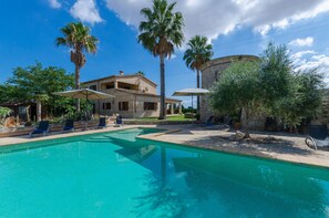 Piscina com espreguiçadeiras, palmeiras e um edifício com fachada de pedra.